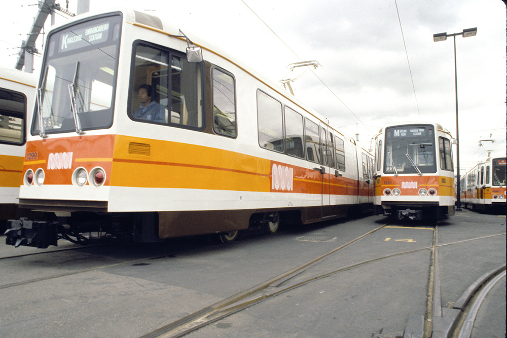 Muni light rail vehicles painted white, orange, and yellow in rail yard.