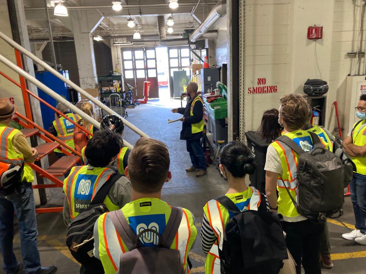 A group of people wearing safety vests and standing among industrial equipment listen to a tour guide who stands in a wide doorway within the Potrero Yard maintenance area.