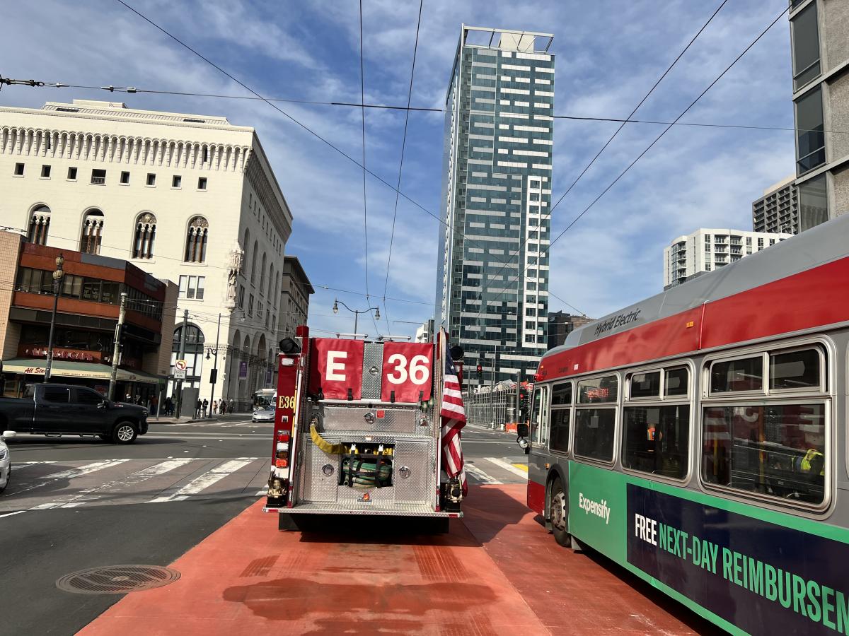 The back of a firetruck is shown passing the side of a Muni bus at the intersection of Van Ness and Market. 