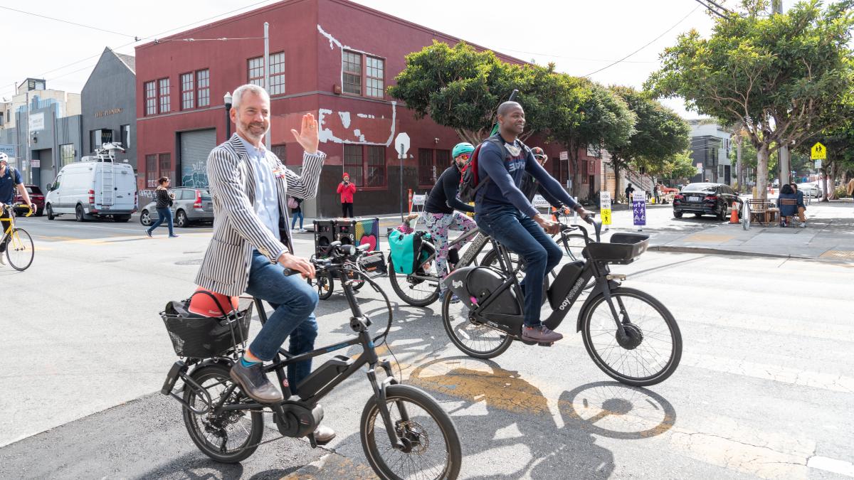 Jeffrey Tumlin riding his bike on San Francisco streets smiling and waving. 