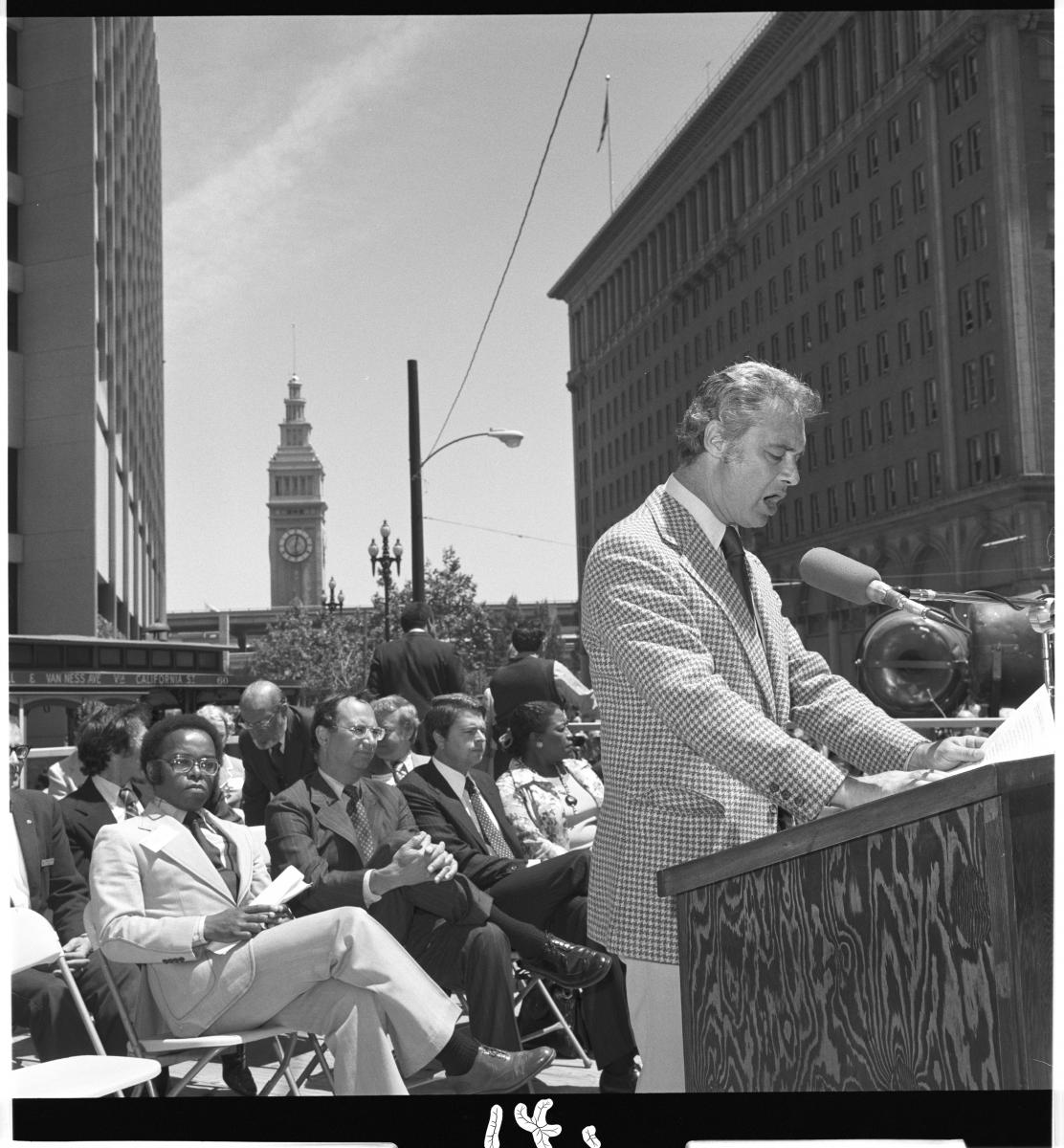 Mayor George Moscone speaking from podium with crowd of people, Ferry Building, and cable car in background