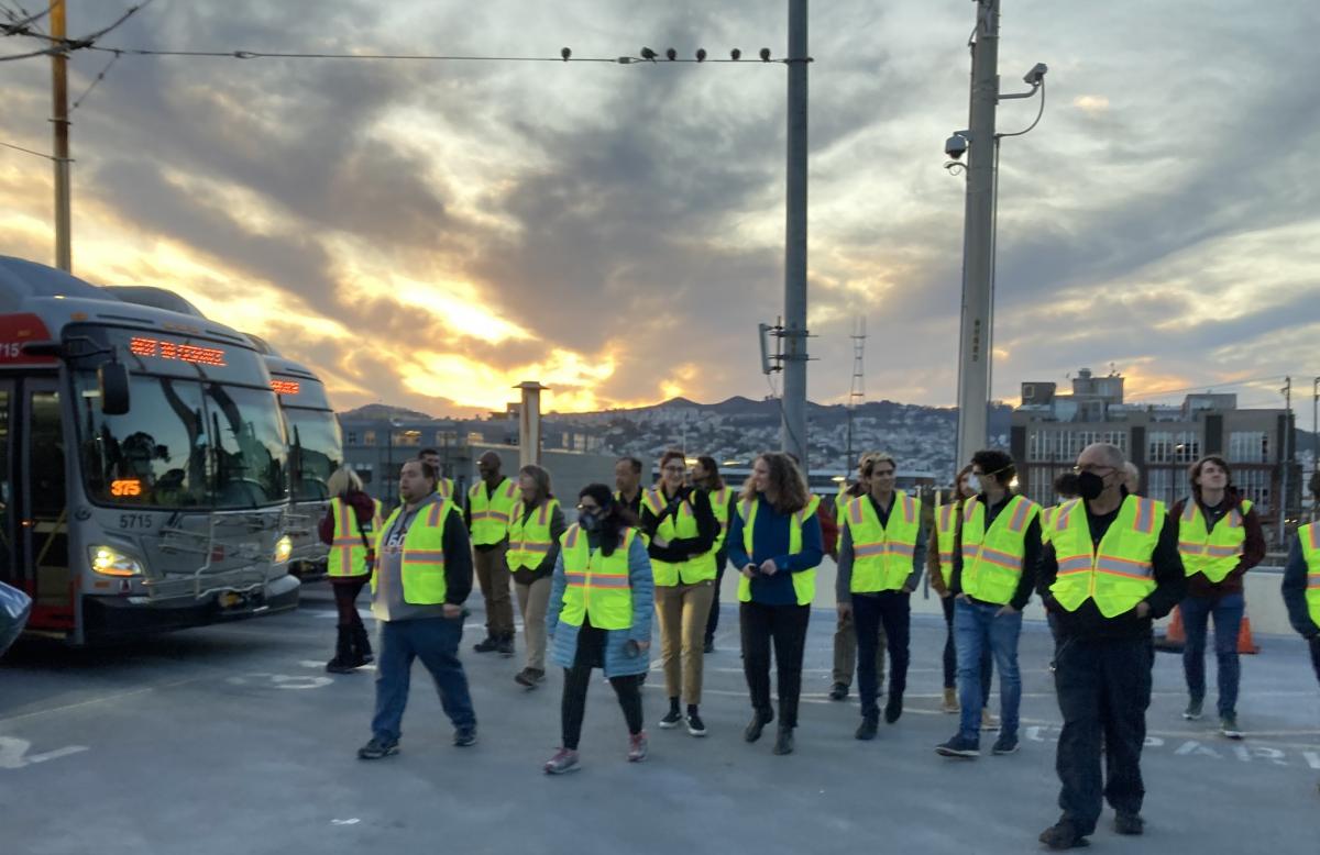 A group of people are pictured walking near buses with a sunset-filled sky in the background.