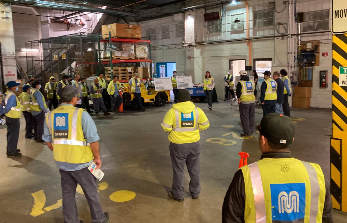 Workers in safety vests are standing inside a bus facility listening to a presenter.