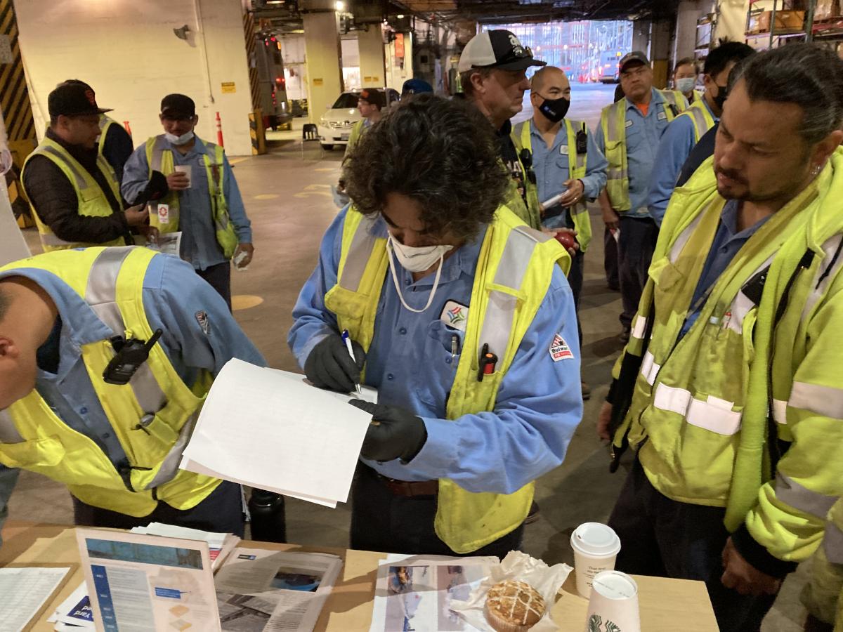 Maintenance workers are standing before a table inside a bus facility and are writing on a clipboard 