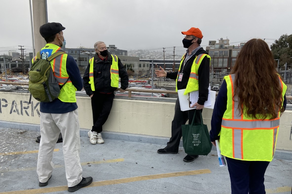 Members from the public and a tour guide wearing an orange hat are seen on the upper level of the Potrero Yard, overlooking buses parked below. 