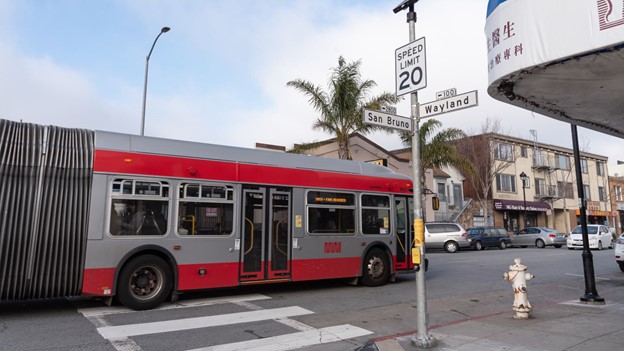 An articulated coach approaching a new 20 mph speed limit sign at the intersection of San Bruno and Wayland Ave.