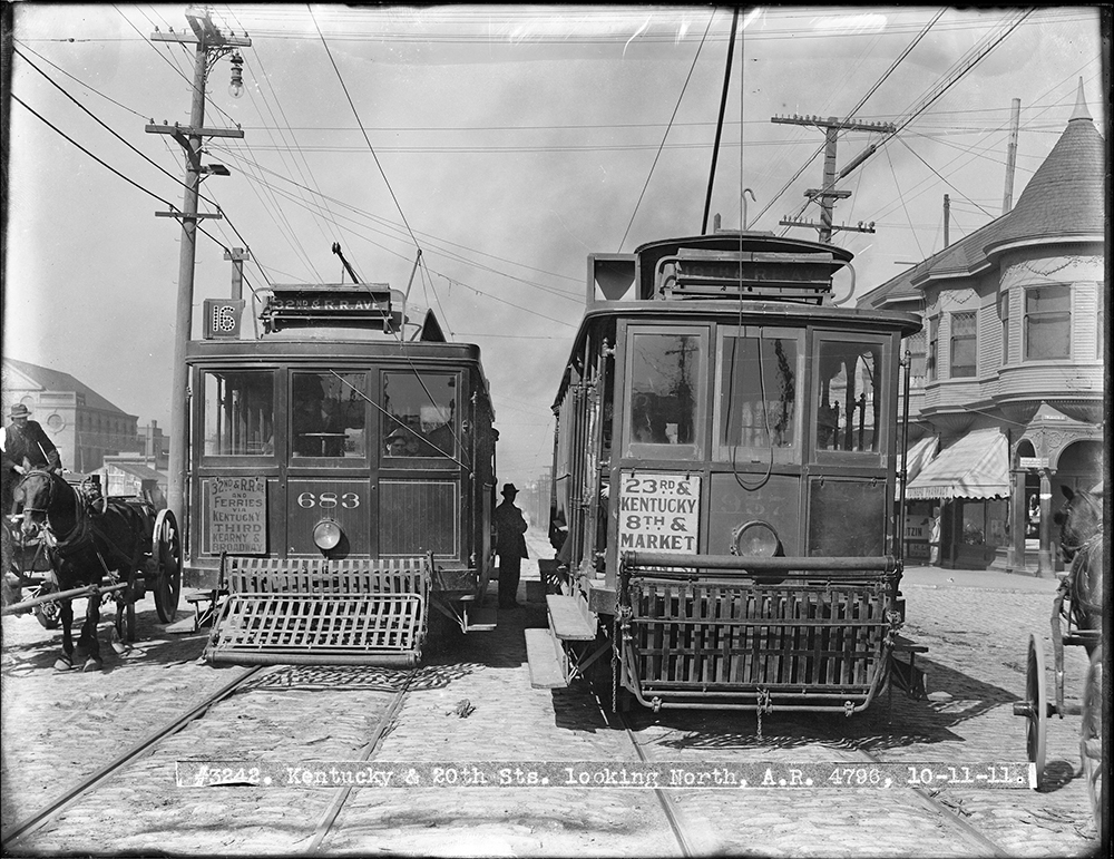 Two streetcars from 1911 shown with a person seen in the middle as well as a horse drawn carriage on the side 