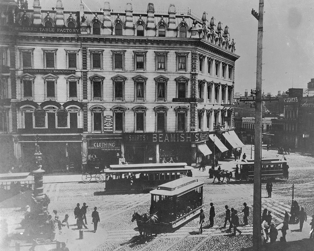 Image of a busy intersection from the 1890's with horse drawn streetcars, pedestrians and a department building are seen 