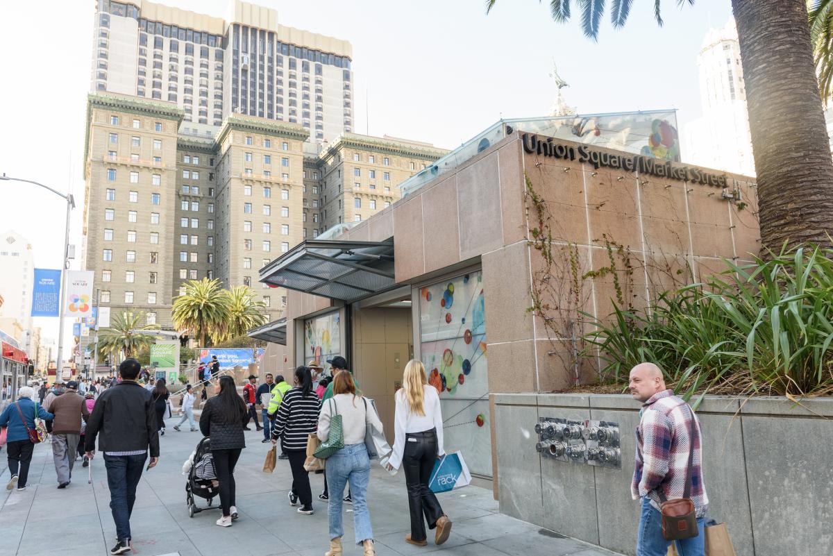 Street view of a station entrance and downtown buildings with dozens of pedestrians holding various items 