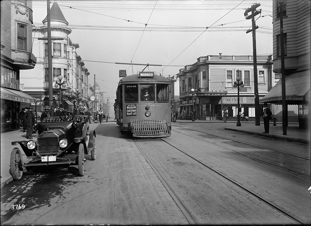 An old streetcar seen on the mainline with a few parked vehicles along the sidewalk and sundry stores in the background