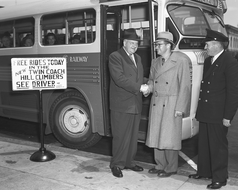 Three people standing next to bus, two people are shaking hands.