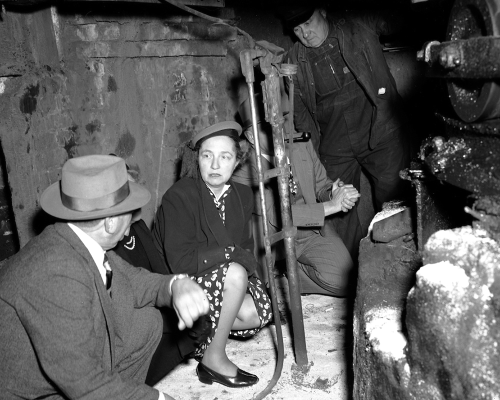 Three people crouching in dirty, small room underneath street with large pulley to run cable car cable