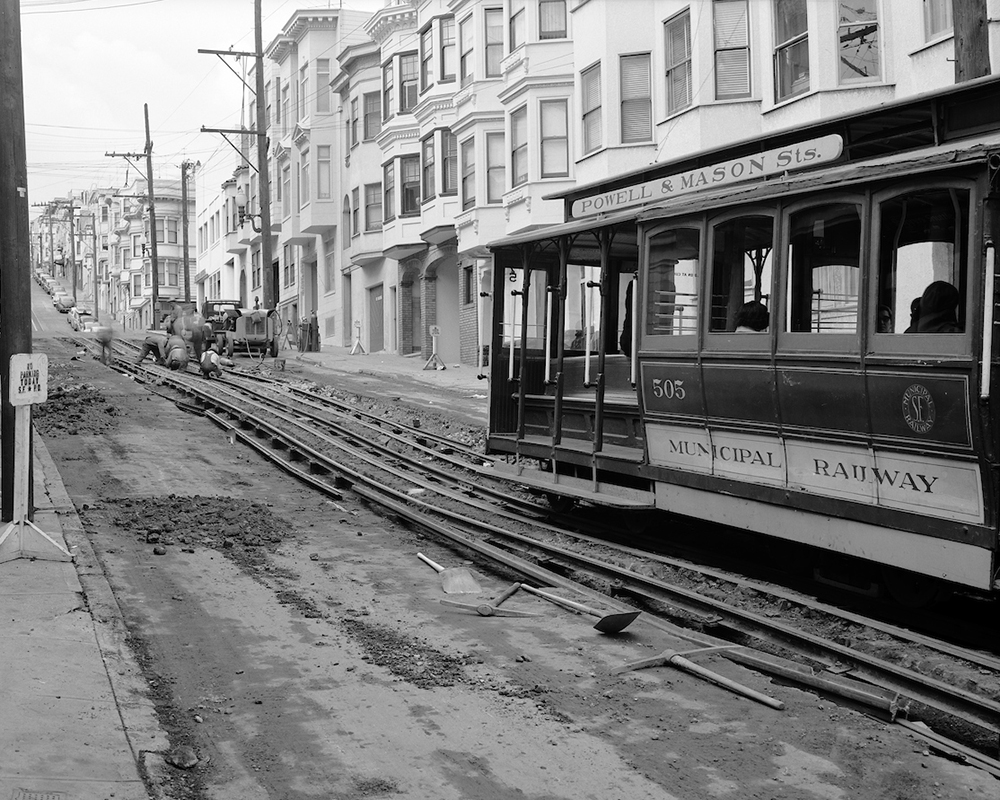 Construction work on cable car line showing exposed tracks and cable car running up street with people working in background