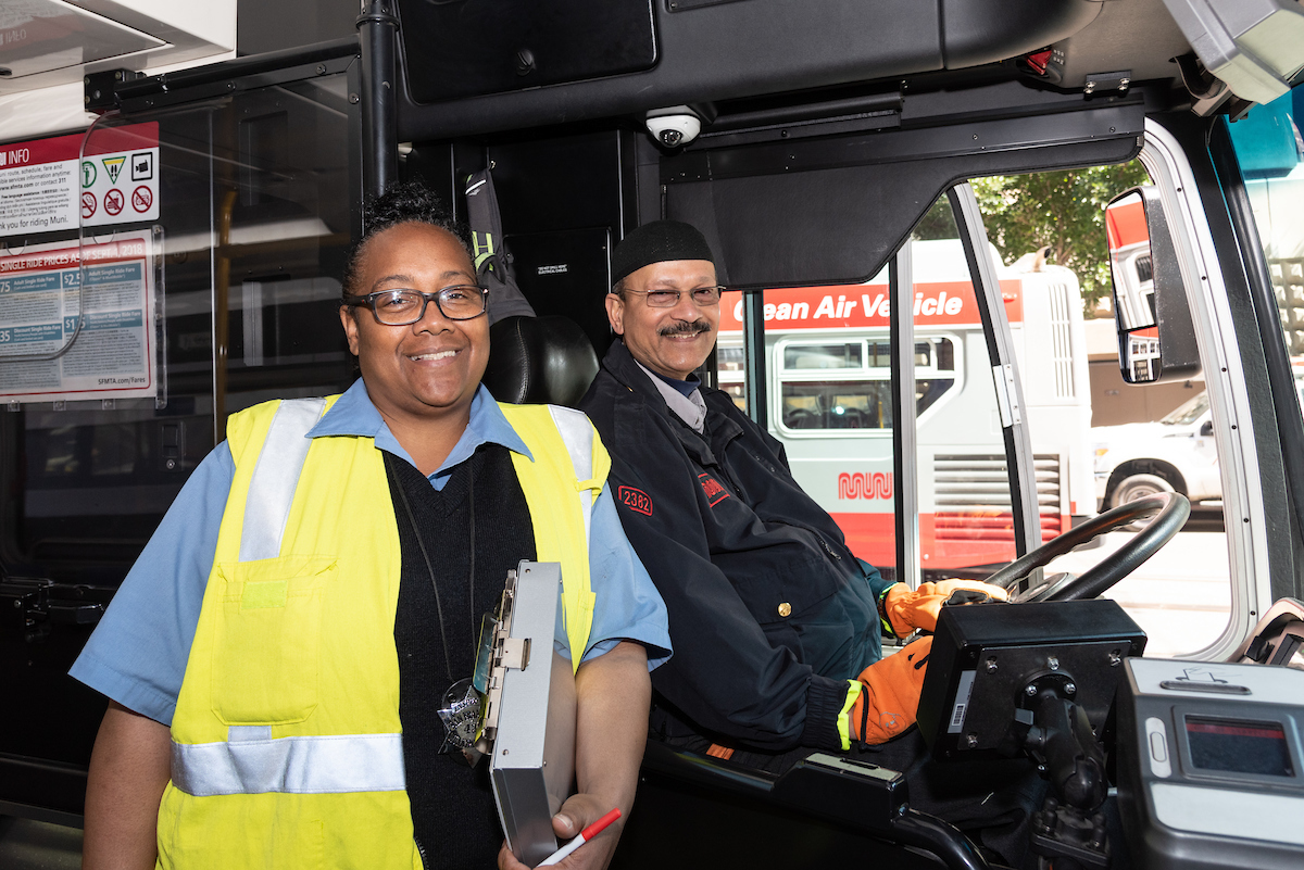 A Muni inspector holds a clipboard next to an operator who's sitting behind the wheel of a Muni vehicle.