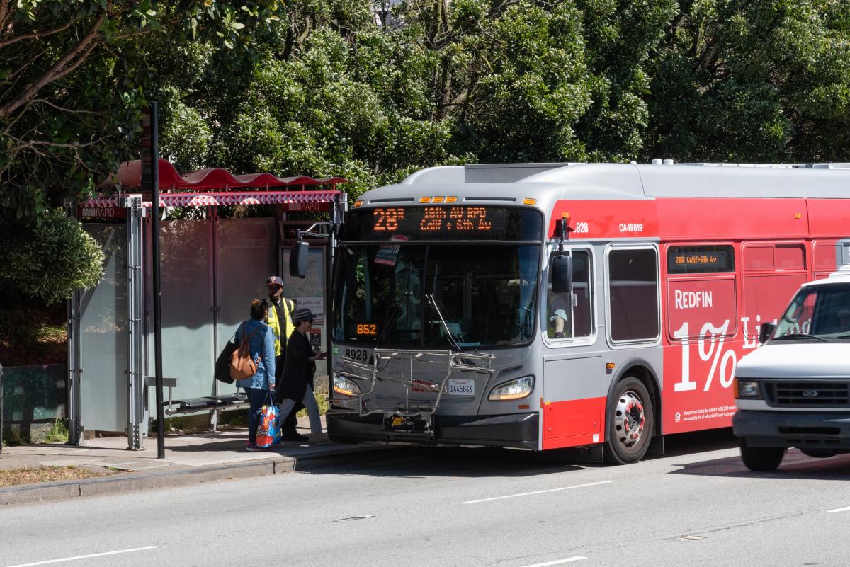 A bus labeled "28R" is pulled over to a bus stop with three people walking onto the bus.