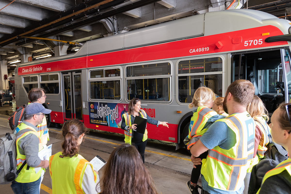 A tour guide standing in front of a Muni bus presents to a group of people inside of Muni’s Potrero Yard facility.