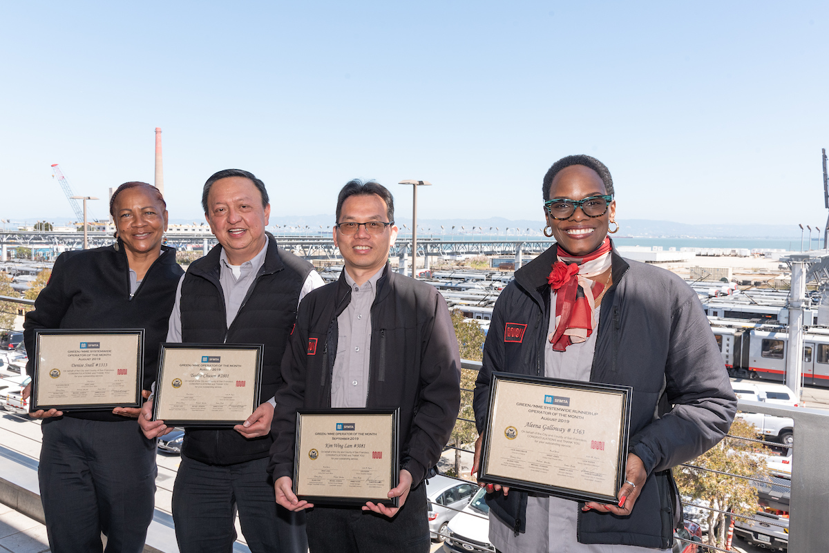 Four Muni operators stand, holding framed certificates with transit vehicles in the distant background.