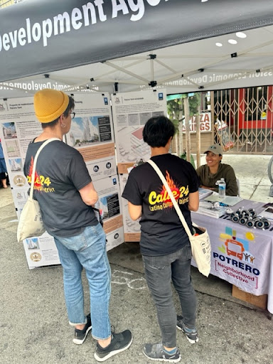 people engage at an outdoor  public event. Visual information materials are spread on a table and are on display in the background on vertical racks. 