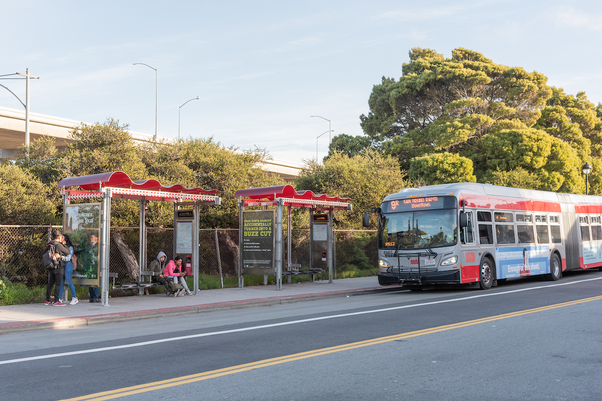 Far shot a bus approaching a bus shelter. There are people sitting and standing. 