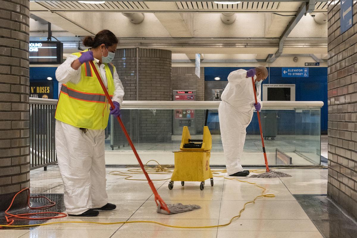 Two Muni custodians wearing masks and yellow vests mop the floor of a station. 