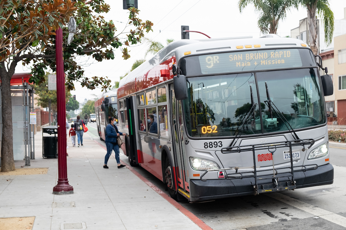 A person is seen boarding the bus along the curb. There are trees in the foreground on the left of the image.