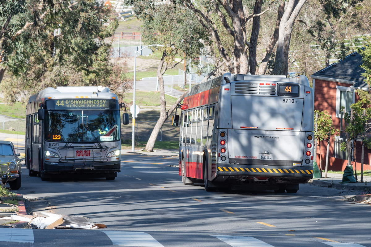 Two buses are side by side traveling in the opposite directions on a street line with trees. 