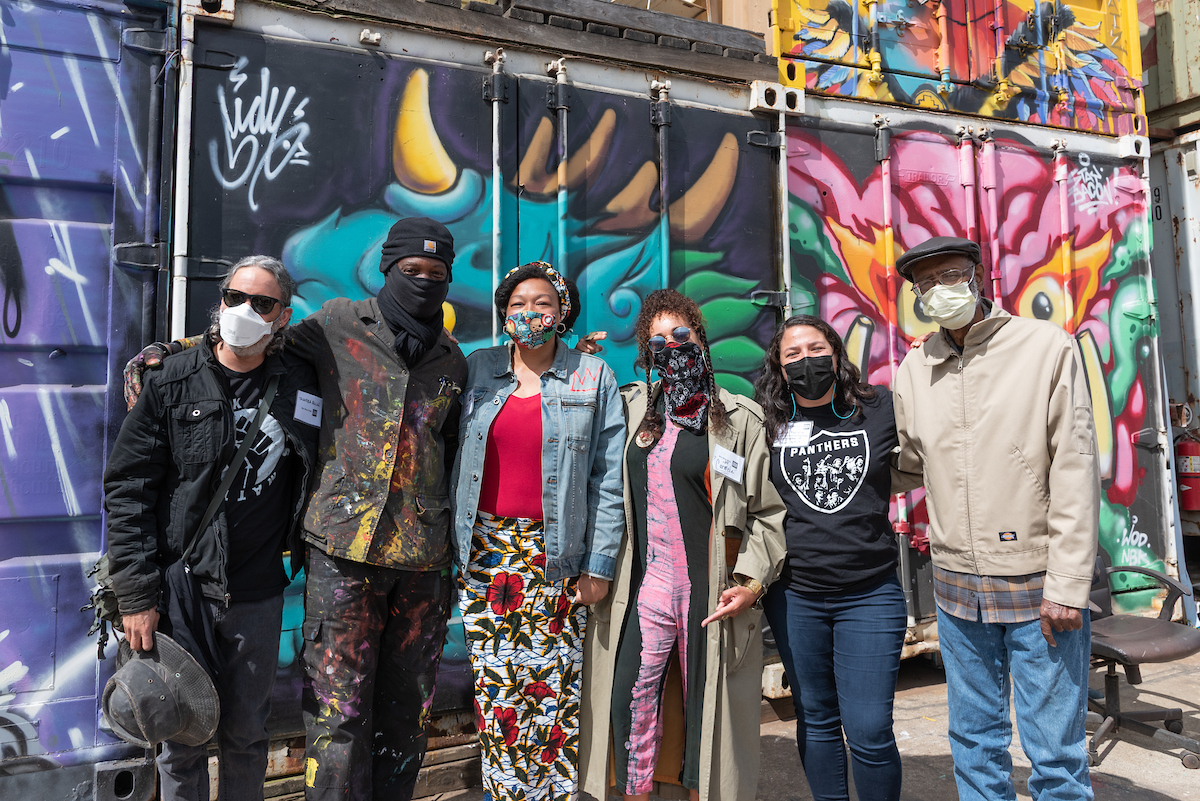 Several individuals with masks huddle for a group photo in front of a colorful mural backdrop in an our setting.