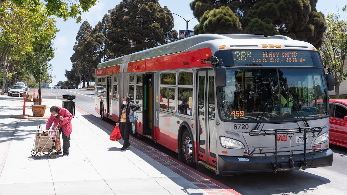 A bus parked on the side of a road with one person exiting with a cart and another person exiting while holding bags