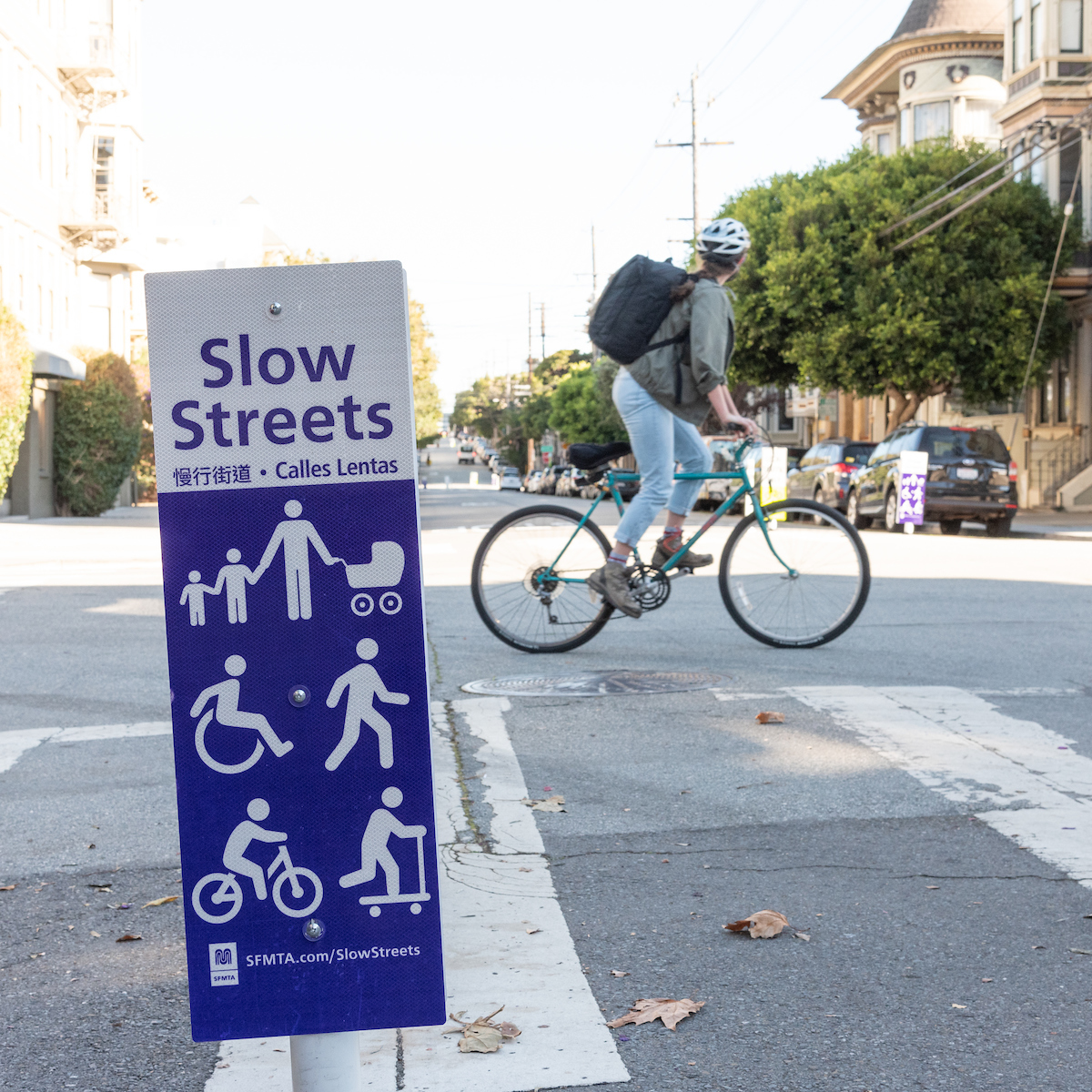 Close up image of a street sign with the words "Slow Streets." Under white figurine wheel users and pedestrians are seen. There is a bicyclist in the background 