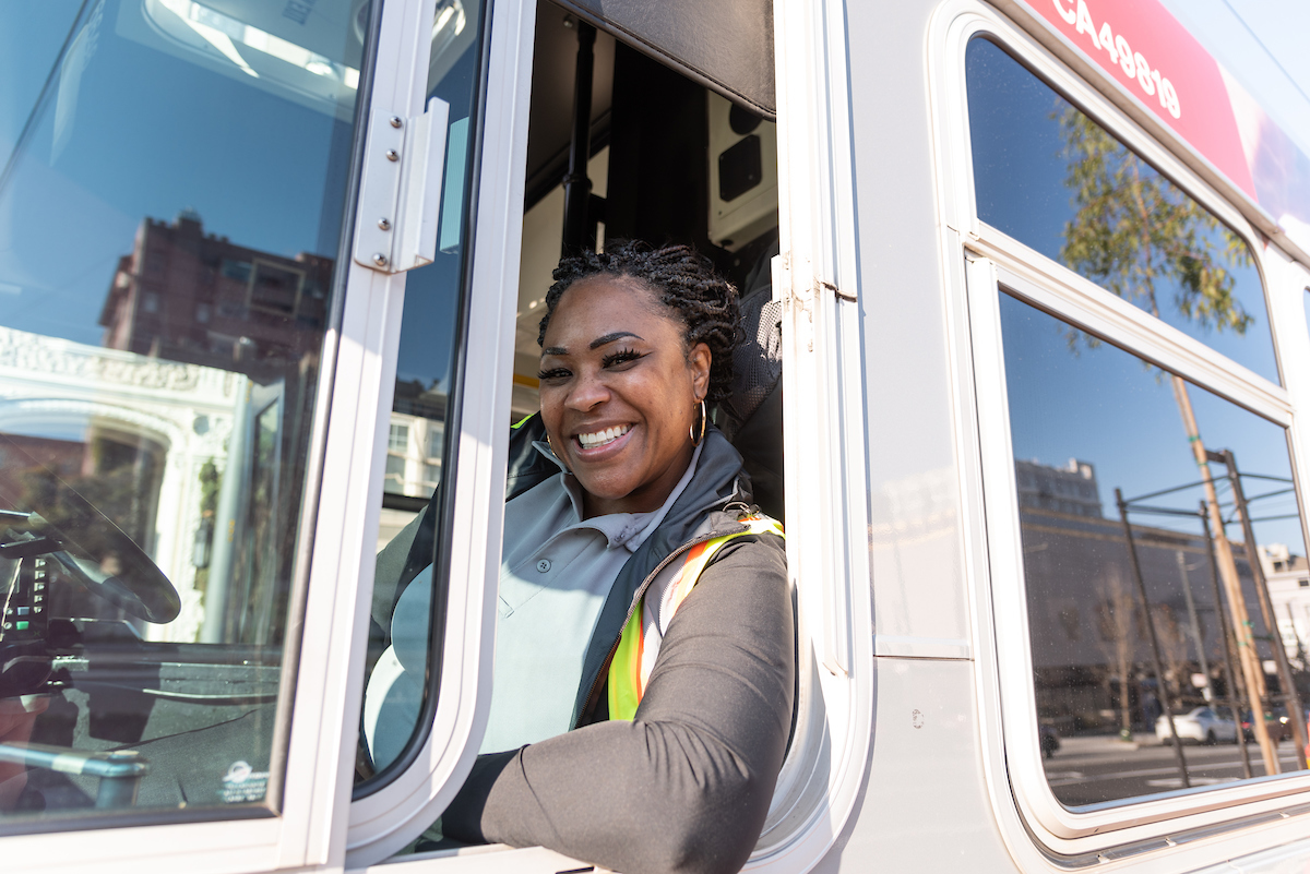 A Muni operator smiles while sitting in the driver seat of a bus.