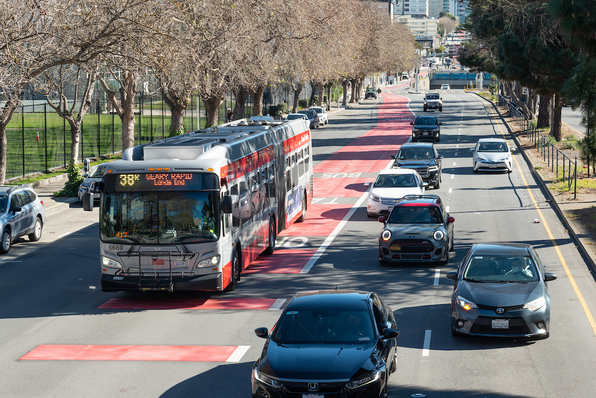 A bus on the left side of the screen is seen on a corridor with over a dozen cars on the right side of the image