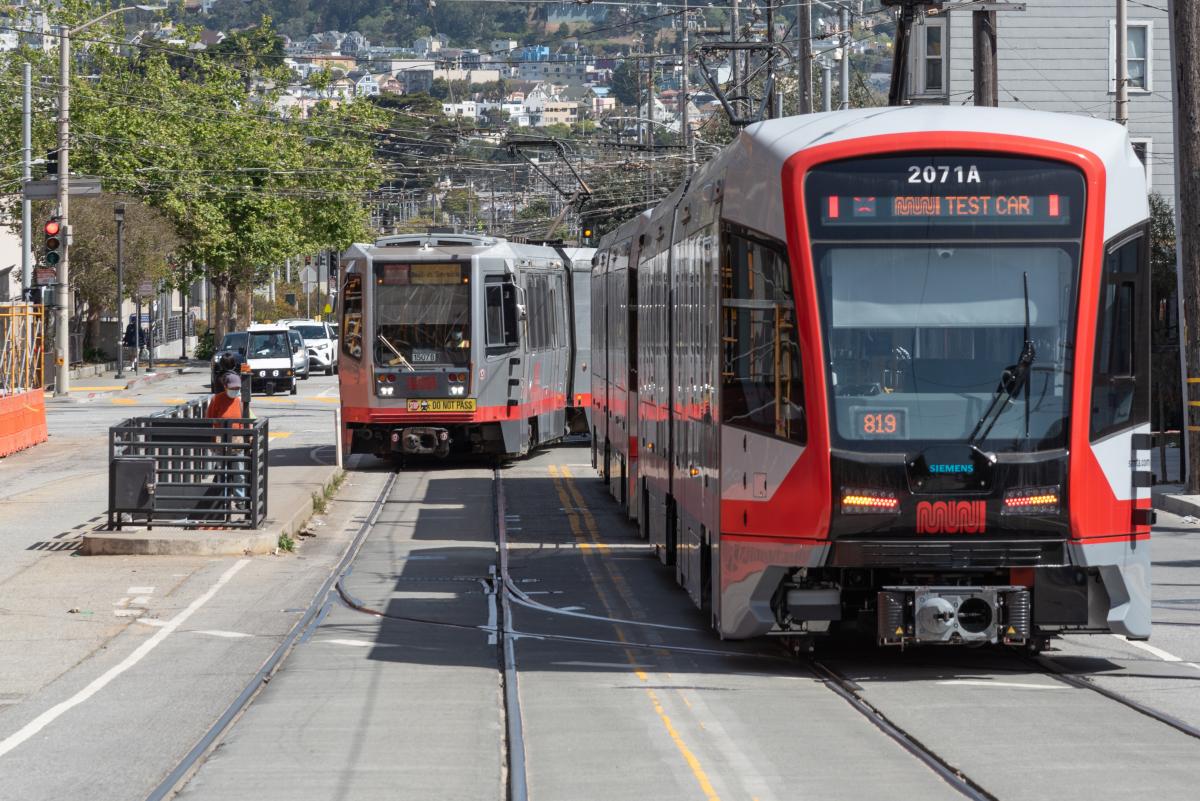 Photo of an older Breda model Muni Metro train on the street in the background, and a new LRV4 model Muni Metro train in the foreground.