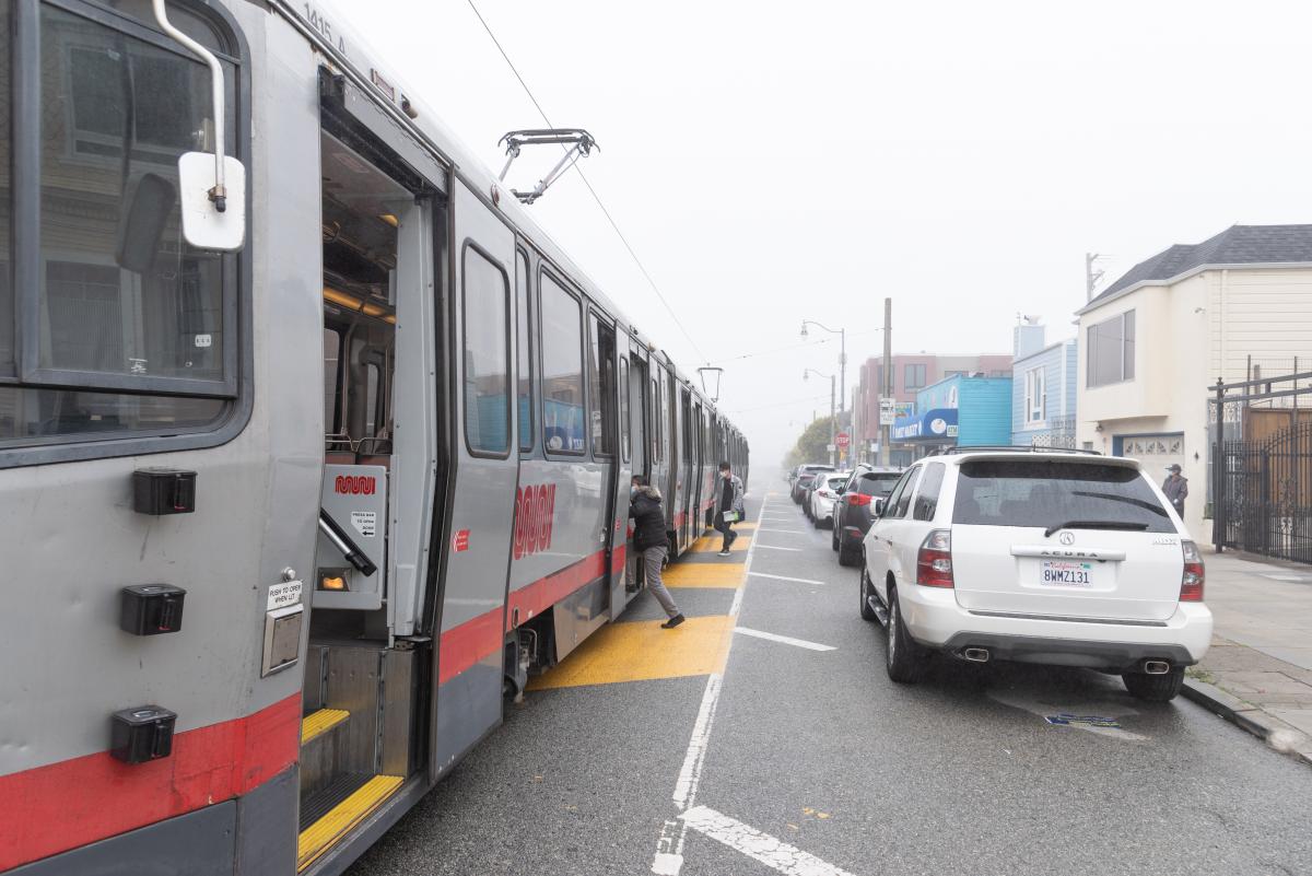 Passengers boarding the train in the street alongside parked cars. 