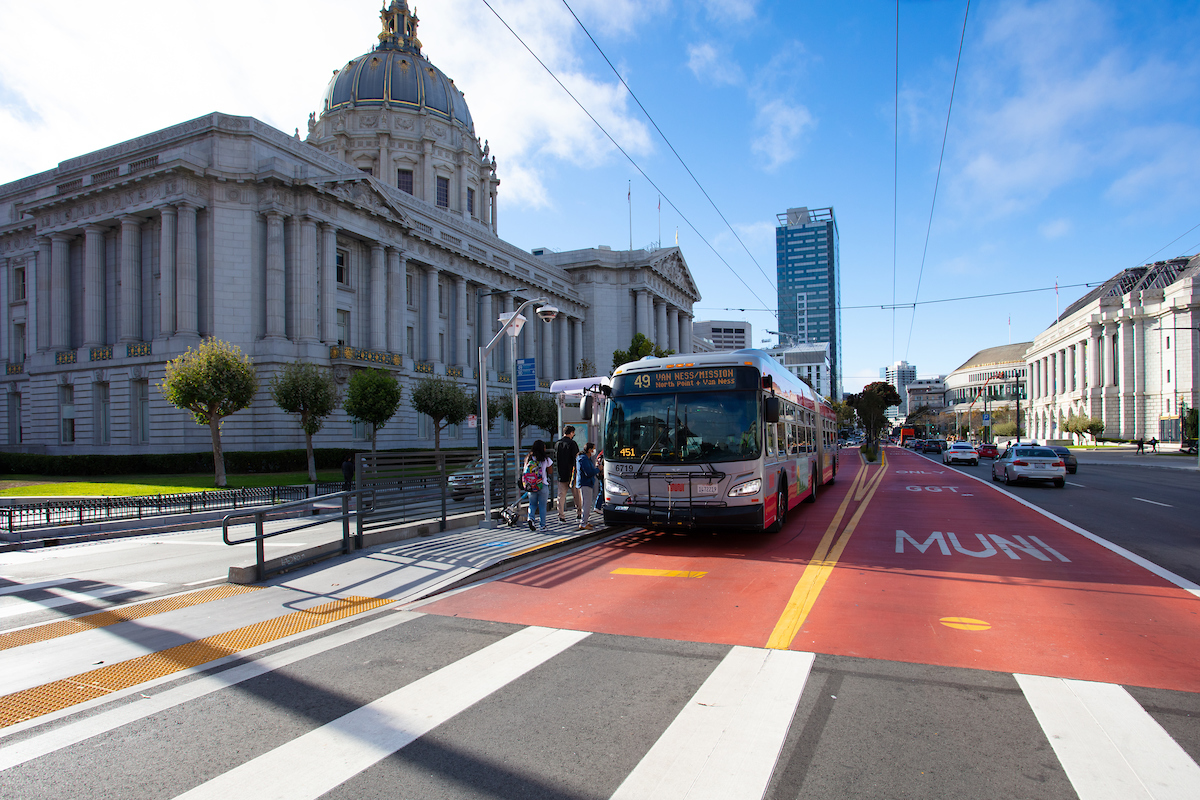 Three people board a Muni bus on Van Ness Avenue in front of City Hall.