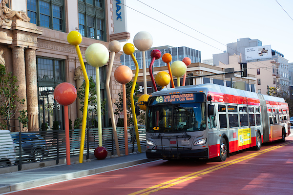 Image features a Muni bus driving down the new red bus rapid transit lane near street sculptures. 