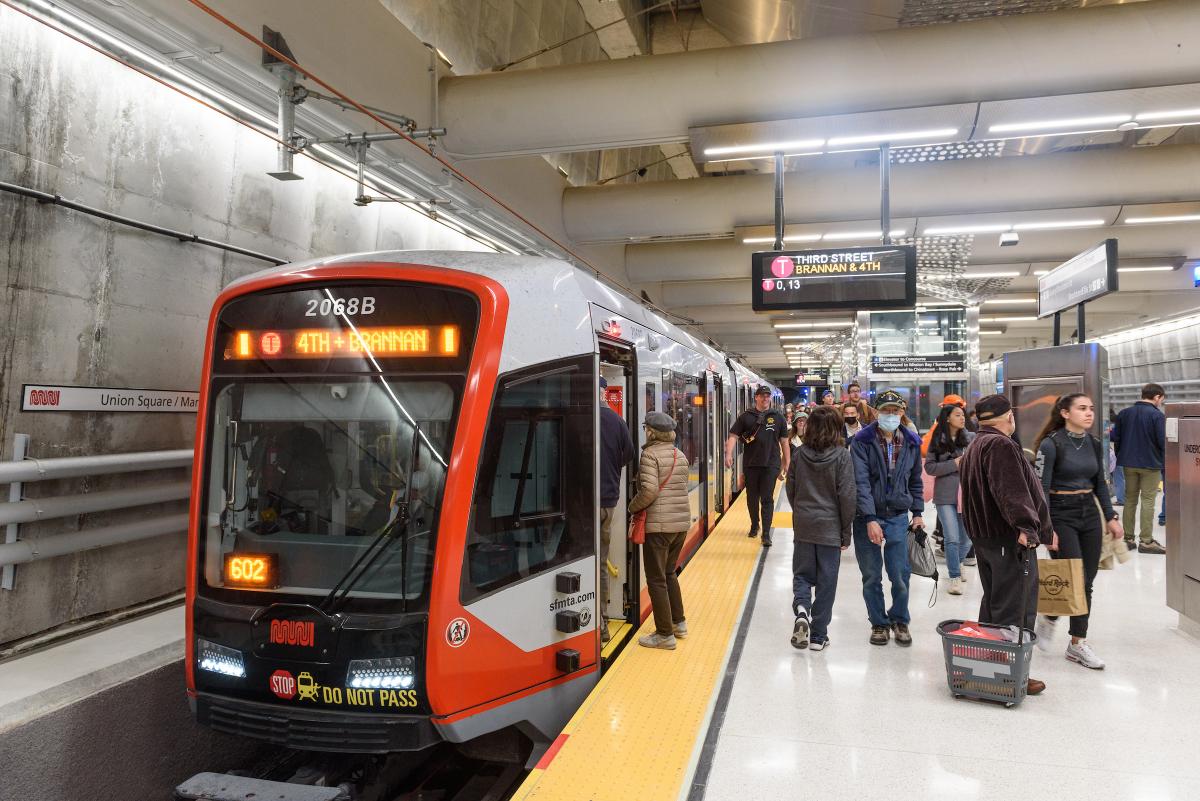 Muni customers boarding the T Third train as it arrives at the station, people walking around.