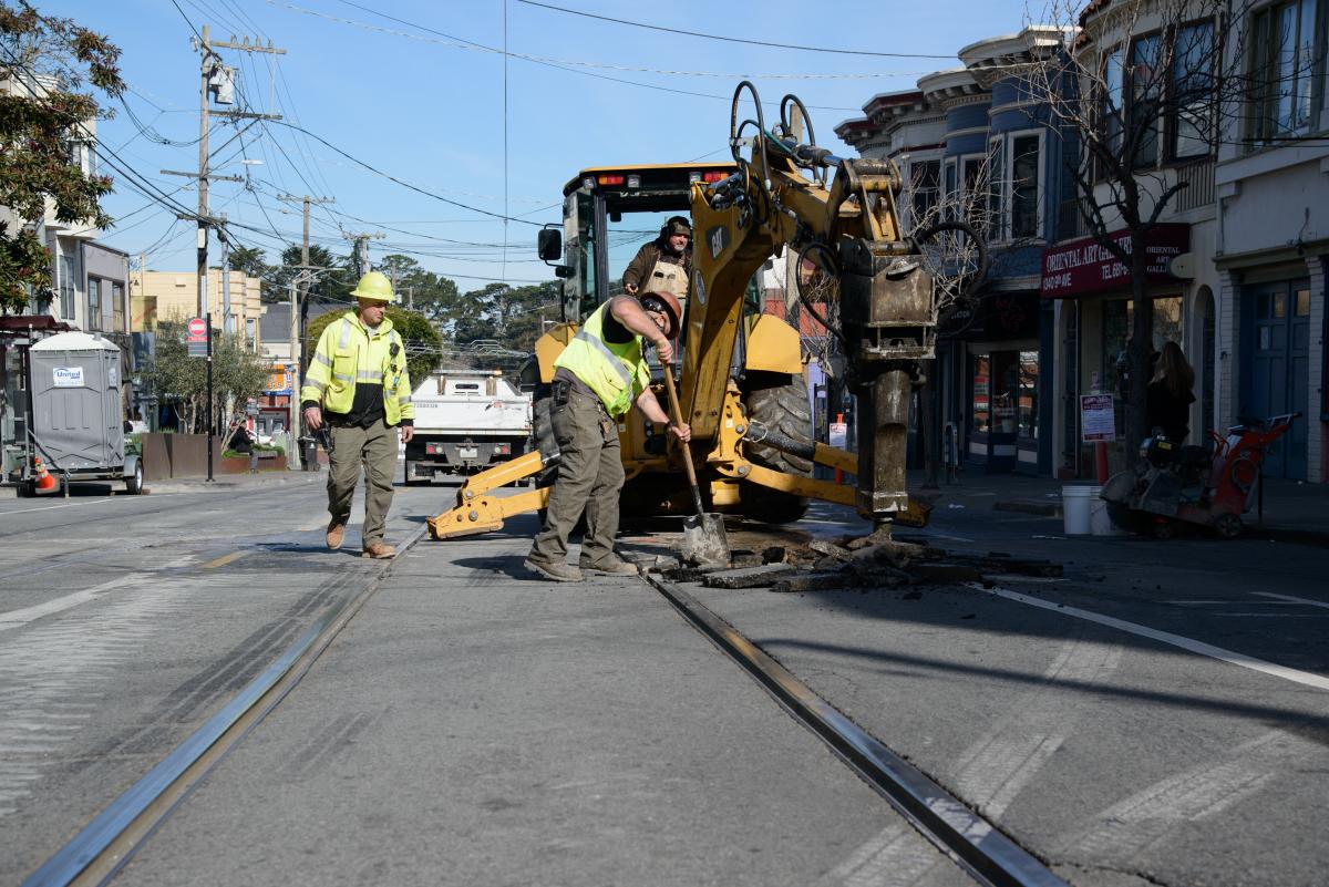 Crew member by the rail track and an excavator with a shovel along the street with houses lined along the side. 