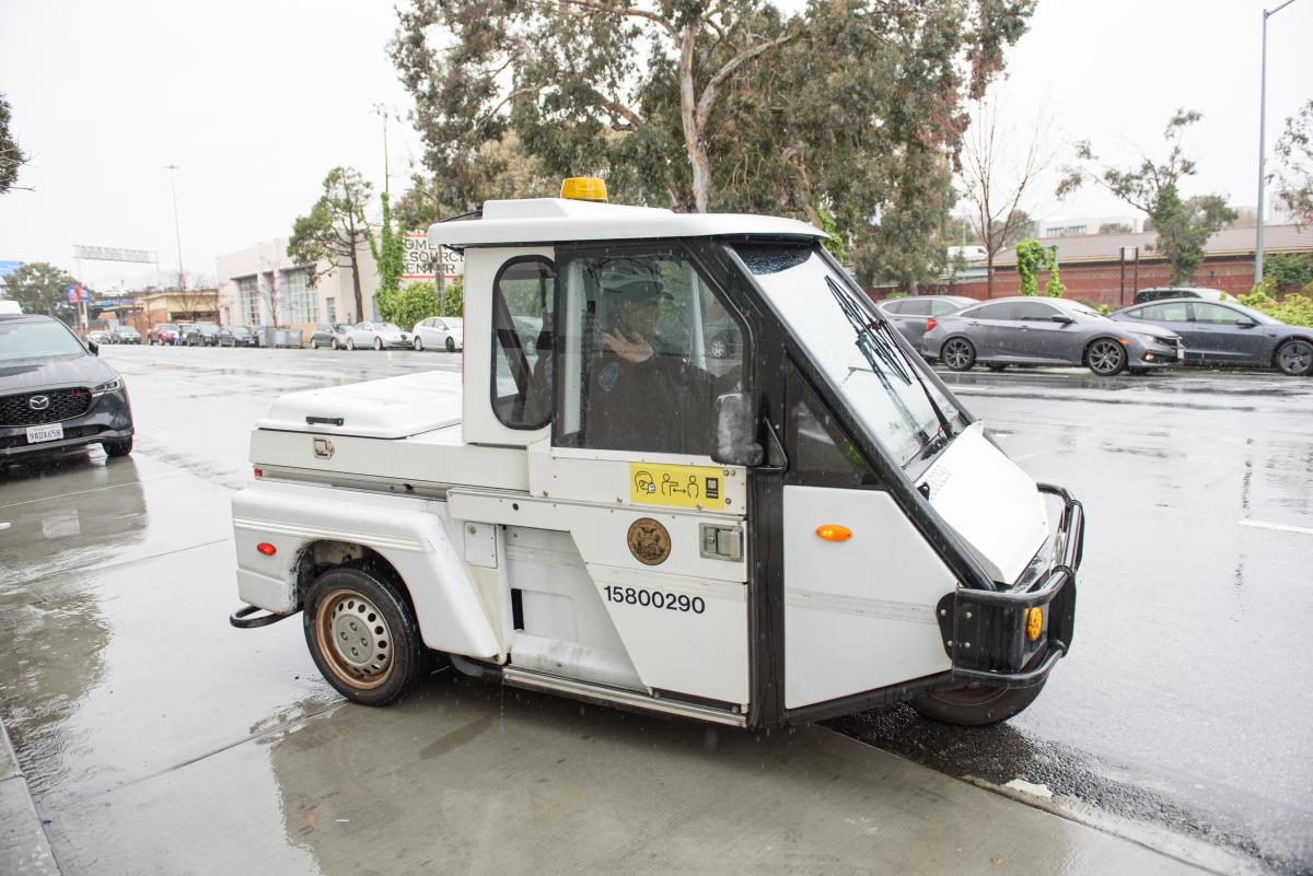 A parking control officer smiles from their vehicle. Rain covers the sidewalk and street near them.