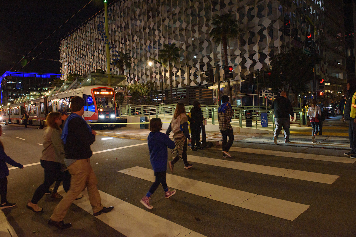 People cross the street to the Chase Center arena in San Francisco at night with a light rail train in the background.