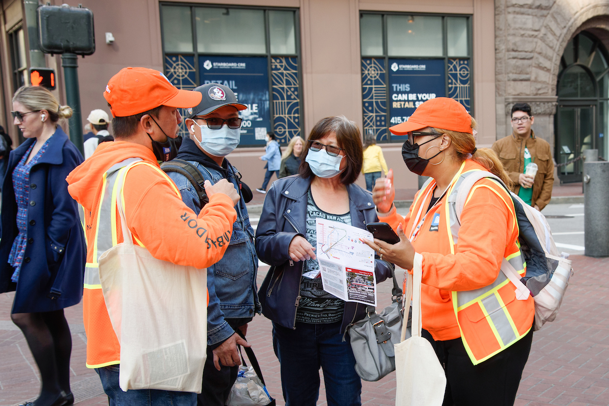 SFMTA ambassadors dressed in orange talk to two people in need of directions.