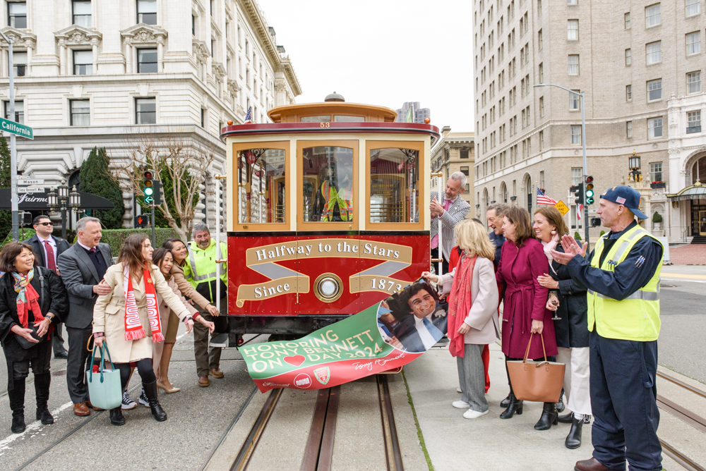 Crowd gathers around Cable Car 53, holding a sign saying the car honors Tony Bennett. The front of the cable car says Halfway to the Stars.