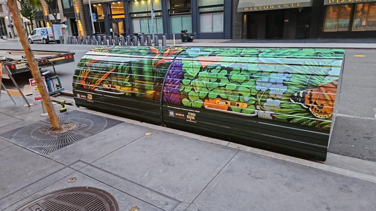 A picture of the new bike hangar storage method by the SFMTA. The hangar is decorated with artwork of plants, and is on the side of the street next to the curb.