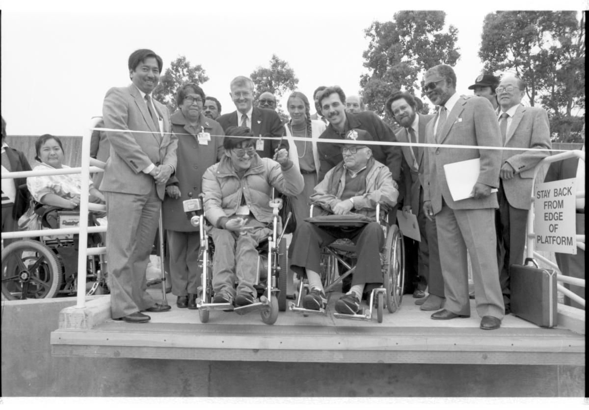 Black and white photo with a crowd of people on an accessibility ramp at a bus stop. Some of the people are standing and some are in wheelchairs. Two people in wheelchairs are in front of a ribbon, one has scissors and smiles as he cuts the ribbon.