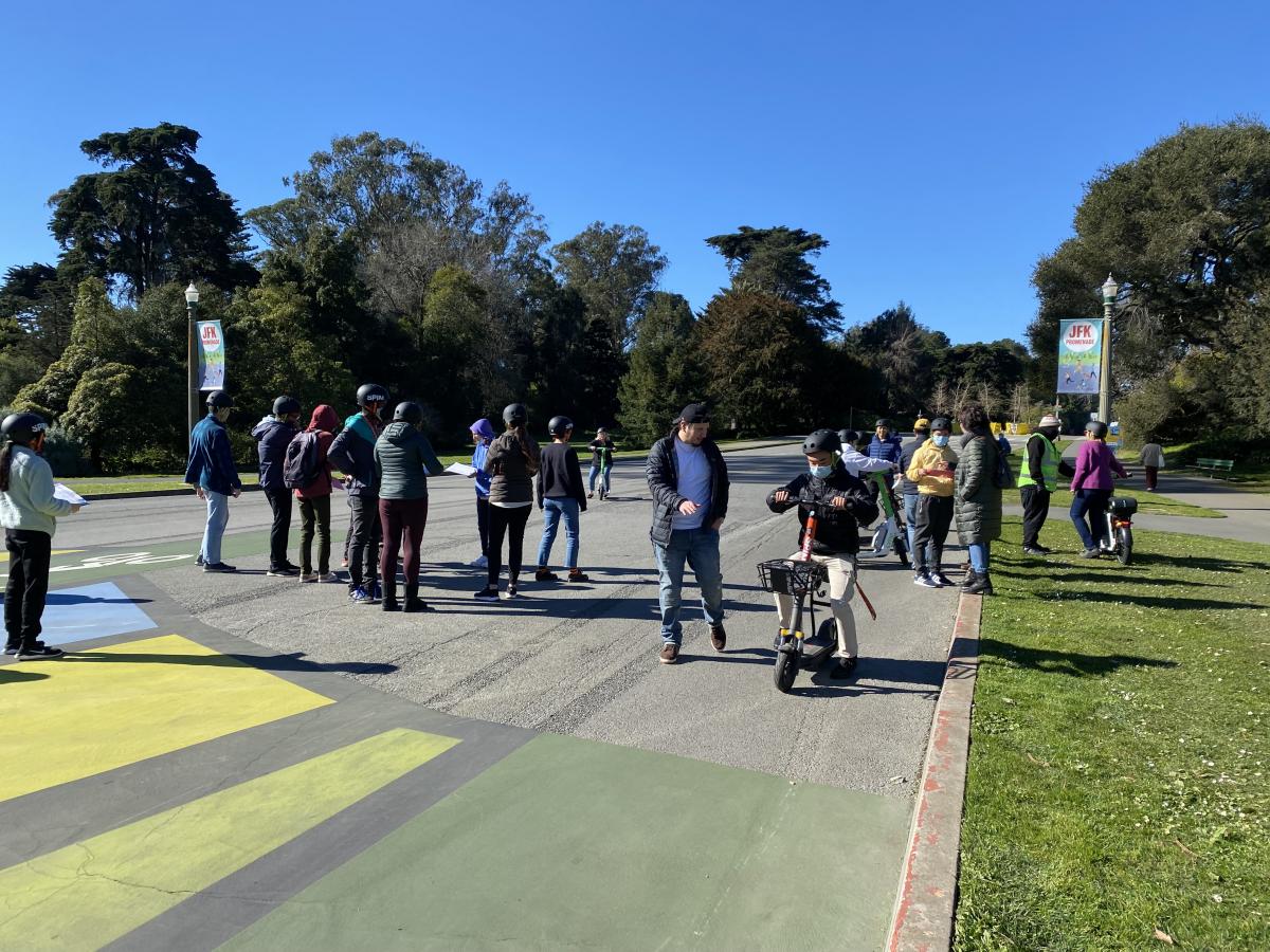 A group of AccessSFUSD students and staff rides adaptive scooters on JFK Drive in Golden Gate Park 