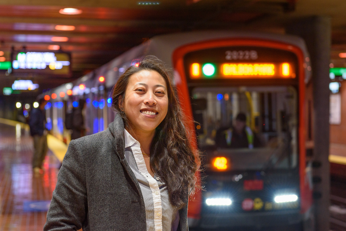 Katherine Kwok smiles on the platform of Castro Station. A train is about to stop in the background.