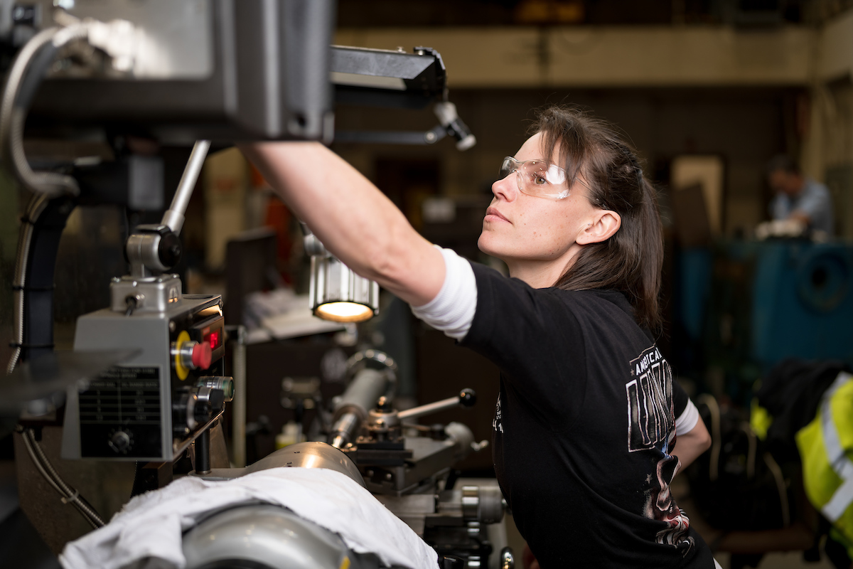 Brittany looks up from a lathe. We see lights and switches near her setup.