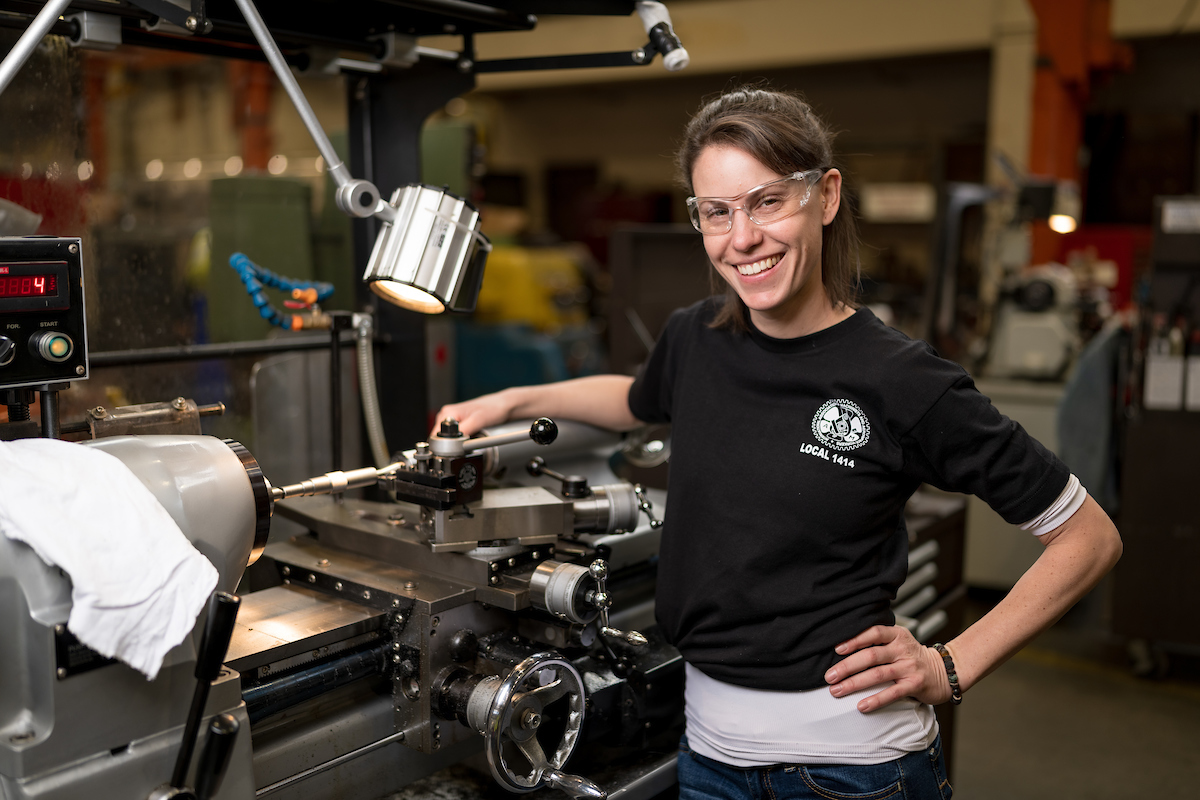 Brittany McMartin smiles over a lathe. We see a range of lights and other machinery around her setup.