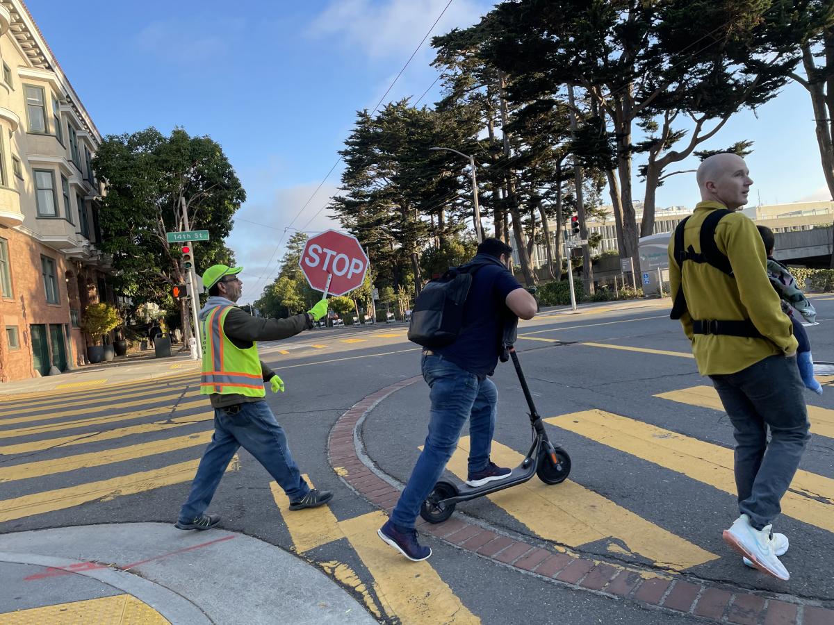 Three people in an intersection. One holding a stop sign, one on a scooter and one with a baby. 