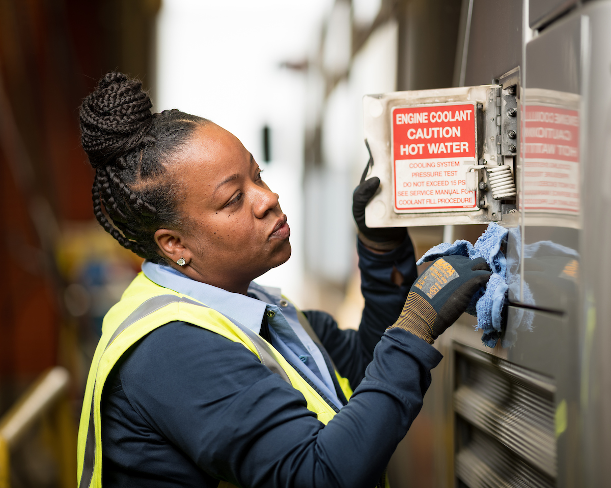 Nicole Humphrey checks the coolant level for a bus. We see her in a yellow vest and blue sweater. She holds a rag as she works.