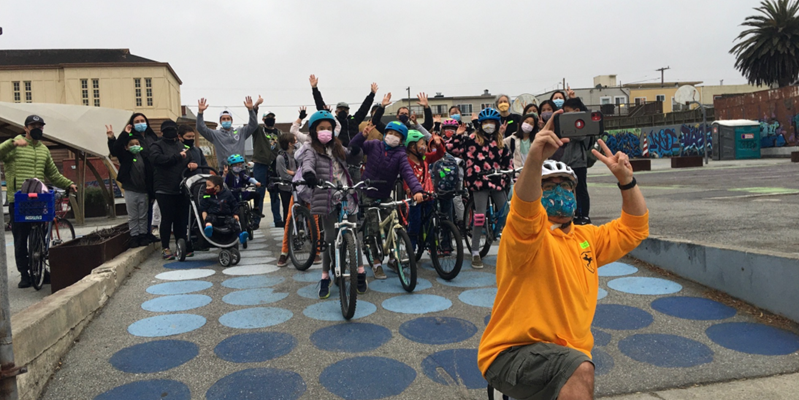 A large group of youth and students taking a photo on a courtyard. Some are on bikes. Everyone is smiling and raising their hands. There is an adult in the foreground taking the selfie picture. 